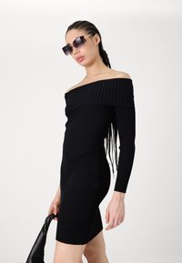 Vestido preto de malha canelada, com ombros à mostra e mangas longas, corte ajustado e tecido texturizado. Modelo com óculos de sol oversized.
