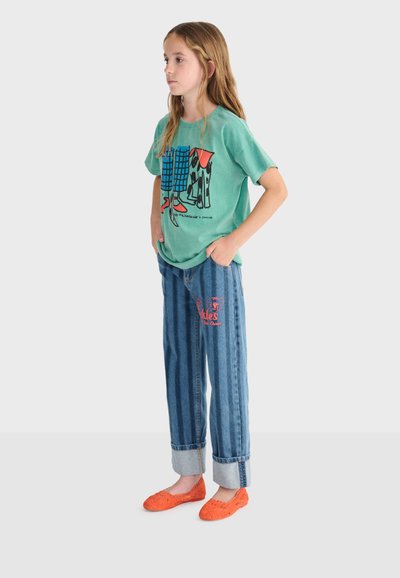 Mlada djevojka stoji sa rukama u džepovima, nosi zelenu majicu s grafičkim printom, plave traperice s preklopima i crvenim natpisom, te narančaste balerinke.