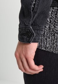 Gros plan d'une main près d'un poignet de veste en jean noire avec un bouton pression noir et un pull en maille gris en dessous.