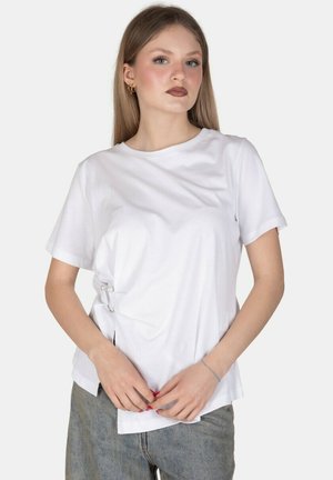 Giovane donna con capelli lunghi che indossa una camicia bianca a maniche corte e jeans, in piedi con le mani giunte davanti a uno sfondo semplice.