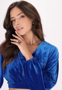 Top corto in velluto blu con maniche a sbuffo, scollo a V profondo e dettagli arricciati. Una collana dorata e un anello completano il look.