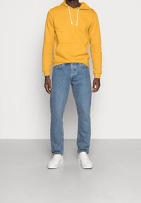 Žlutá mikina s kapucí a bílými šňůrkami, doplněná světle modrými džínami a bílými teniskami, s volným střihem a ležérním designem.