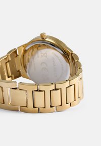 Relógio em aço inoxidável dourado com uma pulseira em corrente. Caixa circular com um fundo de vidro transparente. Possui uma coroa na lateral para ajustar a hora.