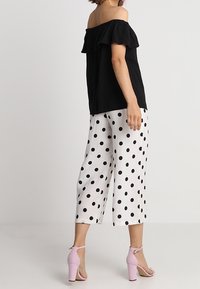 Top nero con spalle scoperte e scollatura arricciata, abbinato a pantaloni bianchi a gamba larga con grandi pois neri. Sandali con tacco rosa.