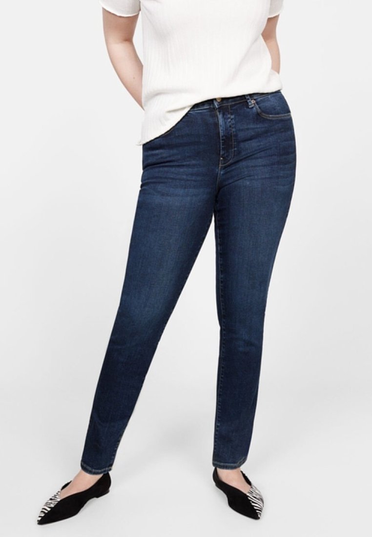 Femme portant un jean skinny bleu foncé, un haut blanc à manches courtes et des ballerines noires pointues avec un motif zèbre sur les orteils, debout les mains dans le dos.
