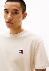 Béžové bavlněné tričko s kulatým výstřihem, na hrudi s malým vyšitým logem "Tommy Jeans" v modré, červené a bílé barvě.