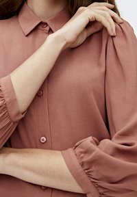 Rose-colored blouse with a soft fabric, buttoned front, and puffed sleeves featuring gathered detailing at the cuffs and shoulders.