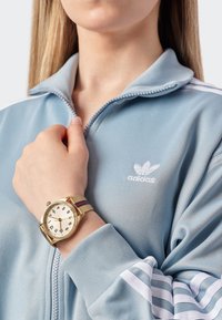 Šviesiai mėlynas zip-up džemperis su baltais raštais ir "adidas" logotipu. Auksinis laikrodis su baltu ciferblatu ir dryžuota medžiaginiam dirželiu. Artimas žvilgsnis į ranką.