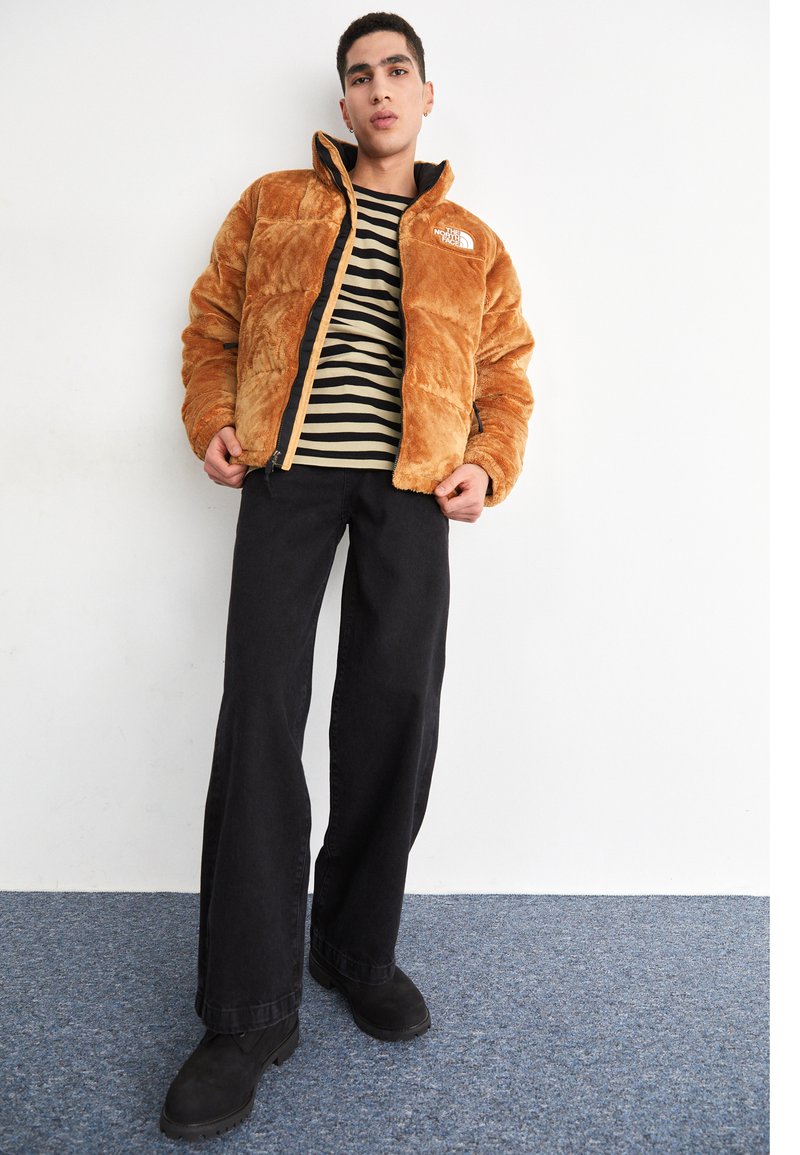 Chaqueta de plumas de imitación de piel marrón con una cremallera negra y logo, combinada con una camiseta de rayas en negro y crema, pantalones anchos negros y botas negras.