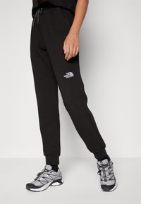Pantalons jogger noirs texturés avec taille élastique, arborant un petit logo blanc. Portés avec des chaussures de sport grises et des chaussettes noires.
