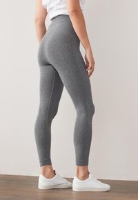 Leggings gris à côtes avec une taille haute, montrant une silhouette ajustée et un motif texturé. Associés à des baskets blanches, debout sur un sol en bois.