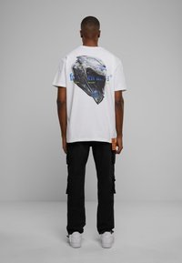 Weißes T-Shirt mit kurzen Ärmeln, das auf der Rückseite ein grafisches Design eines Helms zeigt. Kombiniert mit schwarzen Cargo-Hosen und weißen Sneakers.