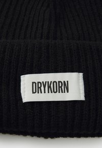 Beanie din tricot negru cu model nervurat, având o etichetă albă care afișează "DRYKORN" în text negru, subliniind materialul texturat și designul simplu.