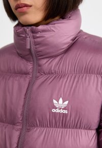 Fialová nafukovacia bunda s vysokým golierom, zipsom vpredu a bielym logom Adidas. Hladká, lesklá tkanina s prešívanou textúrou a stehmi.