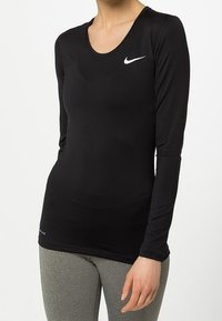 Žena v přiléhavém černém dlouhém rukávu Nike a šedých strukturovaných legínách, stojící s pravou rukou uvolněně podél těla.