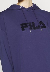 Närbild av en person som bär en marinblå huvtröja med svart FILA-logga och dragsnören synliga vid halsringningen.