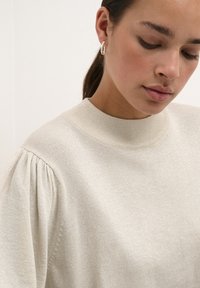 Pulover texturat de culoare bej deschis, cu guler înalt și mâneci puff. Include un detaliu subtil de strălucire, creând o apariție moale și reflexivă.
