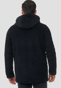 Mann, der eine schwarze, gerippte Cordjacke mit Kapuze trägt, von hinten betrachtet, vor einem schlichten hellgrauen Hintergrund.