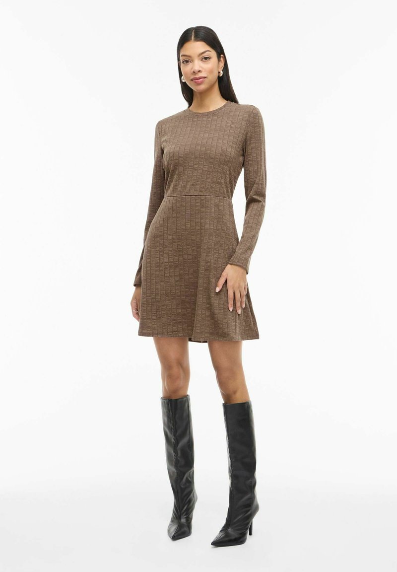 Robe marron à manches longues avec un motif côtelé texturé, une jupe évasée et un col rond. Associée à des bottes noires à talons aiguilles montantes jusqu'aux genoux.