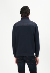 Mørkeblå sweatshirt med strukturert krage og en kontrasterende polstret bakpanel; ribbede mansjetter og hem; kombinert med mørke denim jeans.
