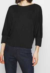 Kvinna iklädd en svart långärmad topp med rynkade manschetter och mörkblå jeans mot en ljus, enfärgad bakgrund.