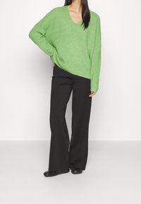 Pull tricoté vert oversize avec col en V, associé à un pantalon noir à jambes larges et des chaussures plates noires. Texture lisse, coupe décontractée.
