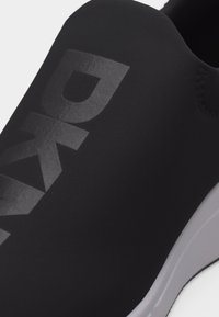Μαύρο αθλητικό παπούτσι τύπου slip-on με μεγάλο ματ λογότυπο DKNY στο πάνω μέρος και λευκή άκρη σόλας ορατή στο πλάι.