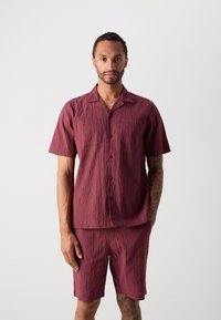 Trumpas rankovės, užsegamas marškiniai ir atitinkamos šortai iš tekstūruoto burgundiško audinio. Pasižymi klasikinio tipo apykakle ir laisvu siluetu.