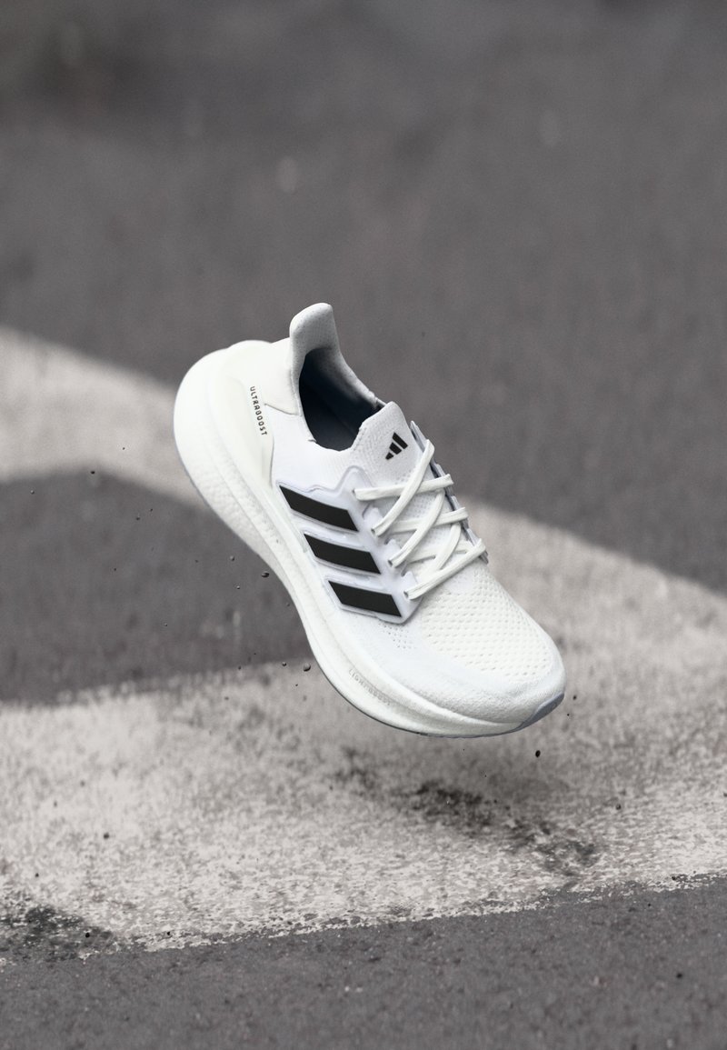 Λευκό παπούτσι τρεξίματος με υφή στην επάνω πλευρά, τρεις μαύρες ρίγες και μαλακή σόλα, τοποθετημένο πάνω σε μια γκρι επιφάνεια με λευκές σημάνσεις.