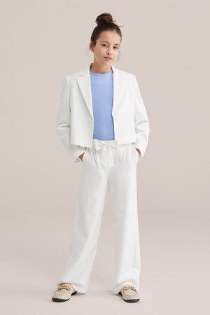 Jauna moteris su susukti plaukais, vilkinti baltą trumpą švarką, baltas plačias kelnes su priekyje surištu diržu, šviesiai mėlyną viršutinį drabužį ir kremine loaferais, stovinti.