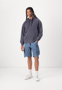 Hanorac oversized, gri, cu șnururi; pantaloni scurți cargo albaștri, cu buzunare; pantofi sport albi cu accente colorate; stând pe un fundal alb.