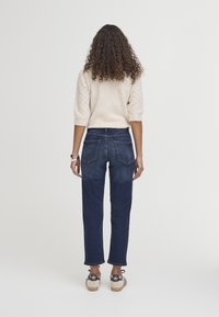 Pull en tricot beige clair avec des manches texturées, associé à un jean en denim bleu foncé avec des poches arrière et un ourlet effiloché, porté avec des baskets.