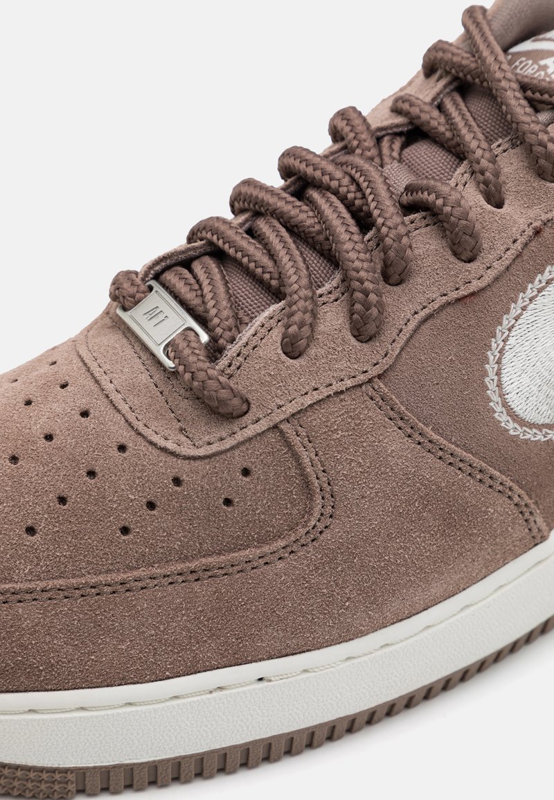 Baskets en daim marron avec une surface texturée, bout rond, lacets tressés, accents matériels argentés et détail de logo blanc sur le côté.