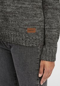 Pull en maille gris foncé avec une finition texturée, comportant un patch logo en cuir. Les poignets et l'ourlet présentent un motif côtelé.