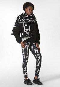 Sudadera de forro polar negra con un bolsillo frontal, combinada con leggings negros que presentan un texto abstracto blanco audaz, y una bufanda a juego con el mismo patrón.