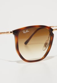 Ochelari de soare Ray-Ban cu un cadru din tortoiseshell întunecat, lentile degradate de culoare maro și accente metalice în ton de aur. Logo-ul este afișat pe lentilă.
