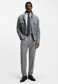 Grauer Wollblazer mit zwei Fronttaschen, hellblau gestreiftes Hemd, gemusterte Krawatte, graue Hose und schwarze Lederschuhe.