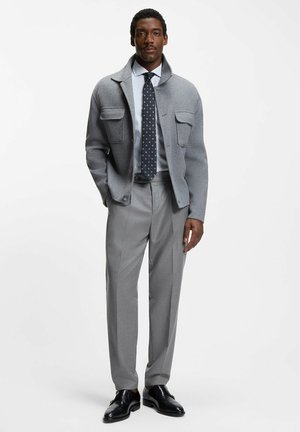 Harmaa villatakki, jossa on kaksi etutaskua, vaaleansiniset raidallinen paita, kuvioitu solmio, harmaat housut ja mustat nahkakengät.