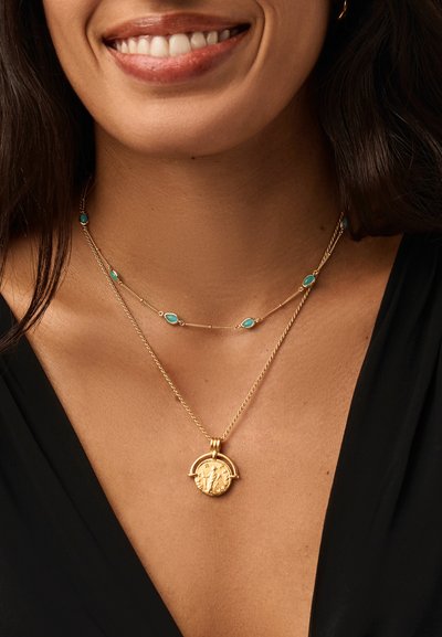 Χαμογελαστή γυναίκα που φοράει δύο χρυσά κολιέ: ένα με μικρές τιρκουάζ πέτρες και ένα άλλο με έναν στρογγυλό χρυσό μενταγιόν, ντυμένη με μαύρο τοπ.