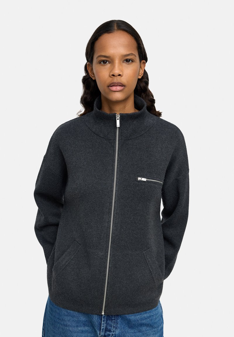 Kobieta o średnio-ciemnej karnacji i falowanych włosach, nosząca ciemnoszary sweter zapinany na zamek z kieszenią na piersi oraz niebieskie dżinsy.