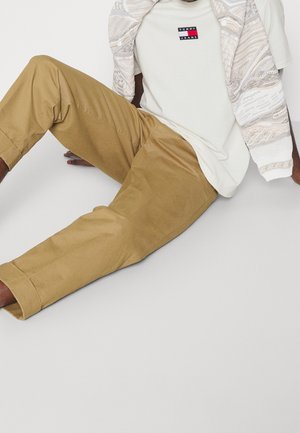 Chino beige avec les poignets retroussés et une coupe décontractée, associé à un t-shirt blanc arborant un patch logo superposé, et un manteau à motif léger.