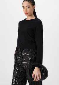 Haut à manches longues noir associé à un pantalon noir à paillettes. Le mannequin tient un sac en cuir noir avec une poignée courbée.