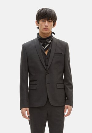 Dunkelgrauer Anzug mit strukturiertem Finish, einreihige Jacke, Reverskragen und passender Weste. Kombiniert mit einem schwarzen Bandana.
