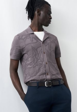 Sötét bőrű, rasztás férfi, mintás szürke rövid ujjú, gombos ingben, fehér alsóing fölött, tengerészkék nadrággal és fekete övvel.