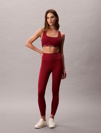 Compleu sport roșu, compus dintr-un sutien sport ajustat cu bandă logo și leggings cu talie înaltă, având text pe lateral. Asortat cu adidași ușori.