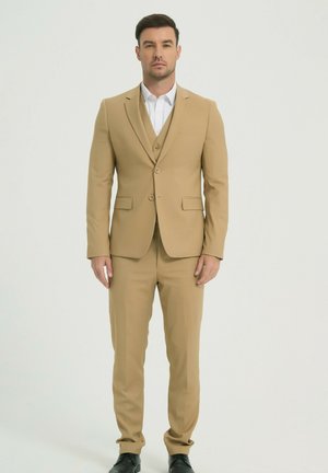 Homme debout, portant un costume trois pièces beige avec une chemise blanche et des chaussures noires, sur un fond clair uni.