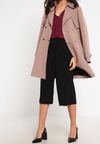 Manteau trench rose clair sur un blouse bordeaux et un pantalon large noir. Des escarpins noirs complètent la tenue. Textures lisses, design structuré.