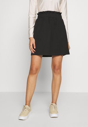 Jupe noire avec taille élastique froncée et poches. Portée avec un haut à manches longues beige et des baskets beiges. Texture lisse.
