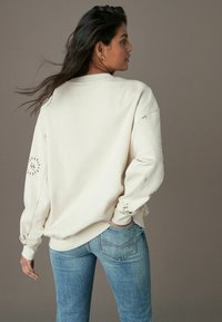 Cream-colored oversized sweatshirt with round neckline and decorative stitching on sleeves. Paired with blue jeans, fitted at the waist.