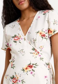 Květinové zavinovací šaty s krátkými rukávy, výstřihem do V, bílým pozadím a řadou růžových, červených a žlutých květin. Hladká textura.